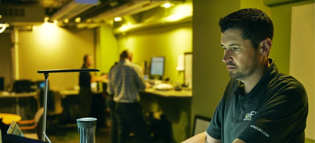 Man sits in office space with many people in background.