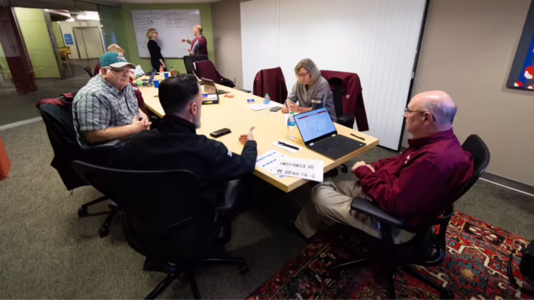 Many people sit in conference room as two people gesture at whiteboard in background