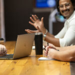 Indian gentlemen sitting at table, gesturing with his hands toward caucasion man seated at the table next to him.