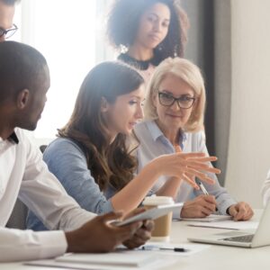 Group of male and female coworkers seated or standing around a female coworker who is gesturing toward a laptop computer.