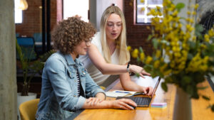 Woman seated in front of a laptop computer with another woman standing over her shoulder gesturing at the computer screen.