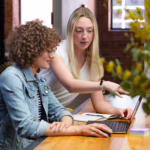 Woman seated in front of a laptop computer with another woman standing over her shoulder gesturing at the computer screen.