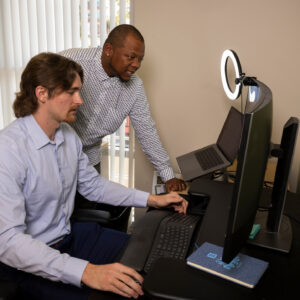 Caucasion man seated in from of a computer screen with African American man standing over his shoulder also looking at the computer screen.