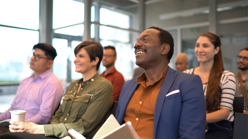 Group of business people sitting in chairs looking forward and smiling as if at a lecture.