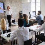 Woman stands at front of class gesturing toward a TV screen.