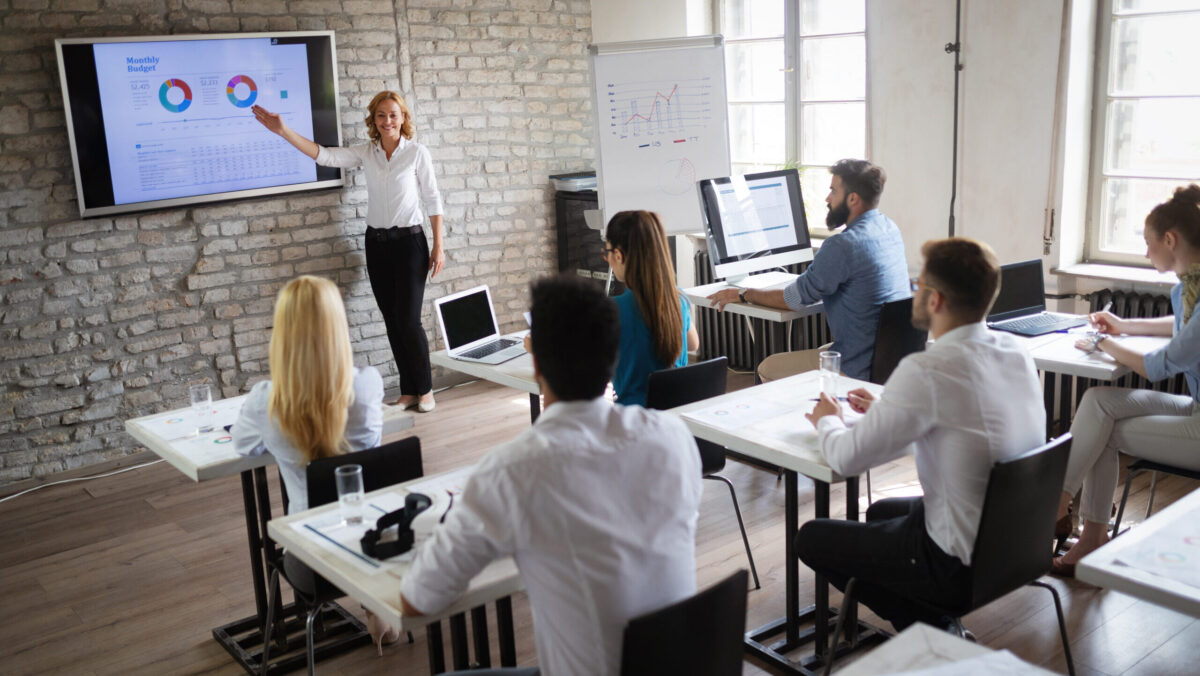 Woman stands at front of class gesturing toward a TV screen.