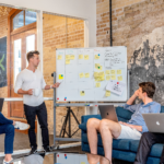 Group of men sitting around a white board with Post-It notes on it having a conversation.