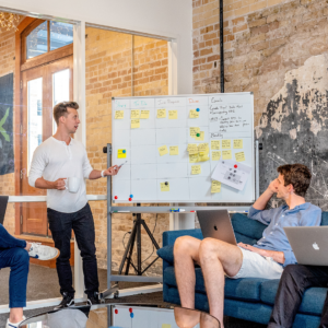 Group of men sitting around a white board with Post-It notes on it having a conversation.