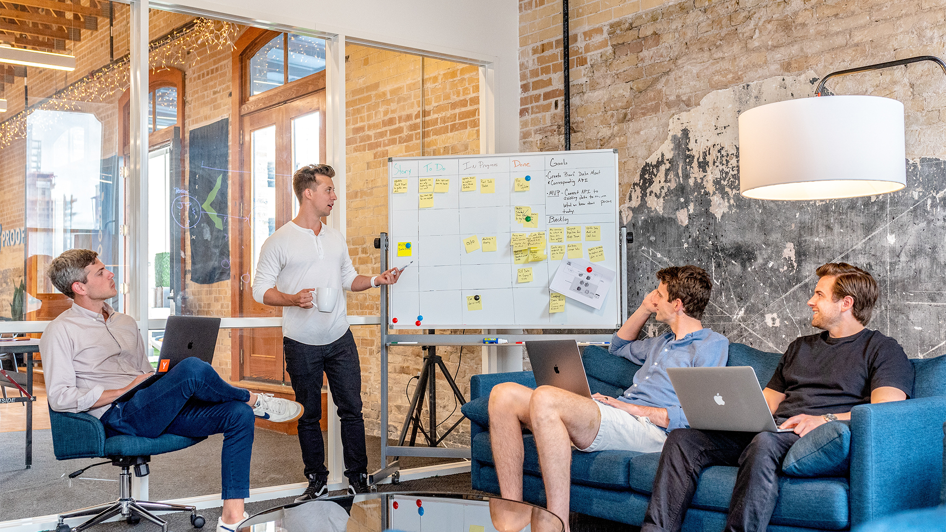 Group of men sitting around a white board with Post-It notes on it having a conversation.