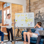 Man standing in front of white board with post-it notes and writing while a group of coworkers look onward.