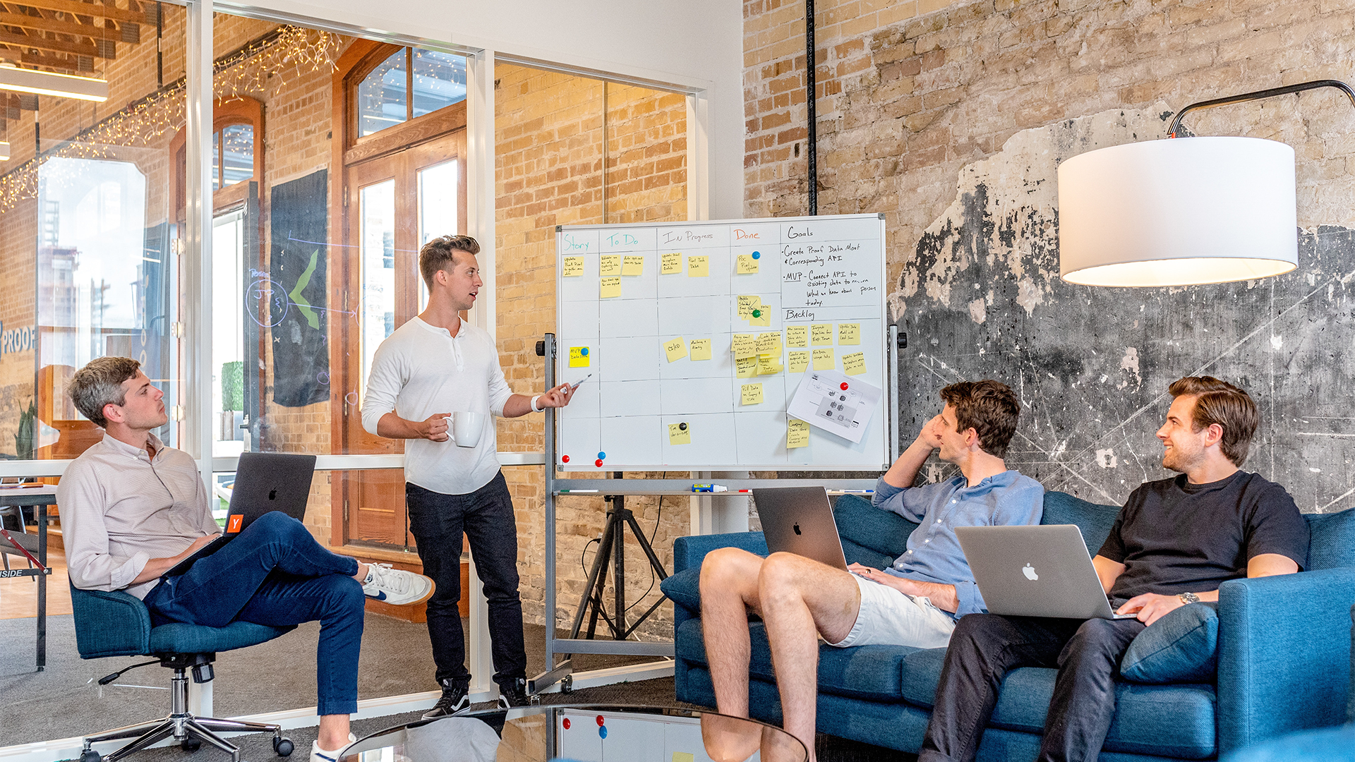 Man standing in front of white board with post-it notes and writing while a group of coworkers look onward.