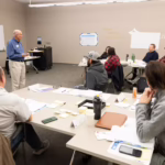 Instructor standing at the front of a classroom of project management students lecturing.