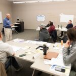 Instructor standing at the front of a classroom of project management students lecturing.