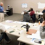Instructor teaching at the front of a classroom with wall charts and post-its hanging on the room walls.