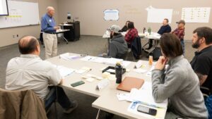 Instructor teaching at the front of a classroom with wall charts and post-its hanging on the room walls.