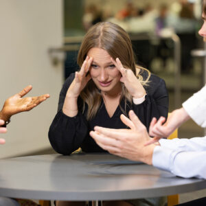 Woman with her hands on her temples listening to people around a table talking and gesturing toward her with their hands.