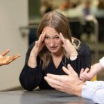 Woman with her hands on her temples listening to people around a table talking and gesturing toward her with their hands.