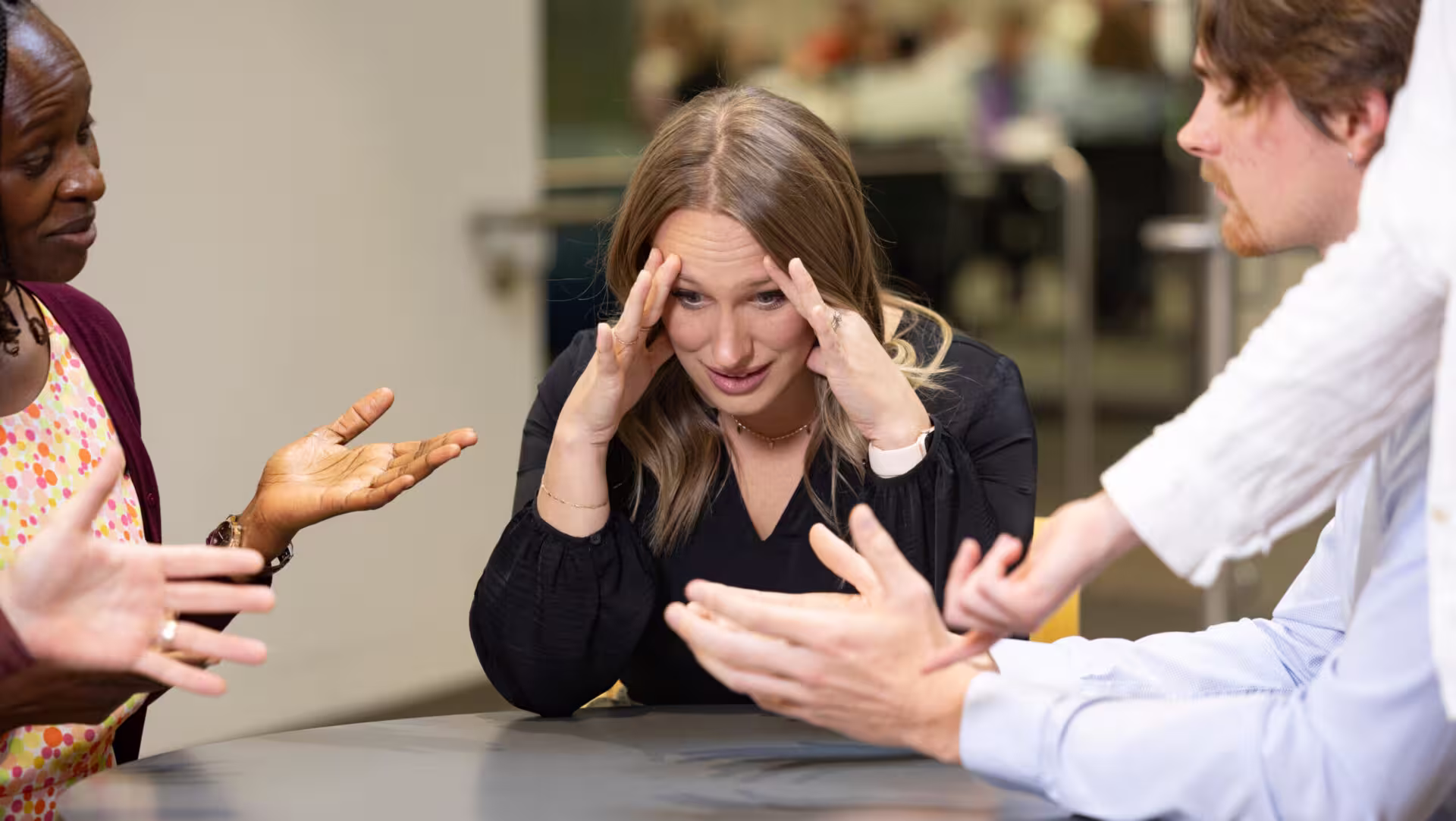 Woman with her hands on her temples listening to people around a table talking and gesturing toward her with their hands.