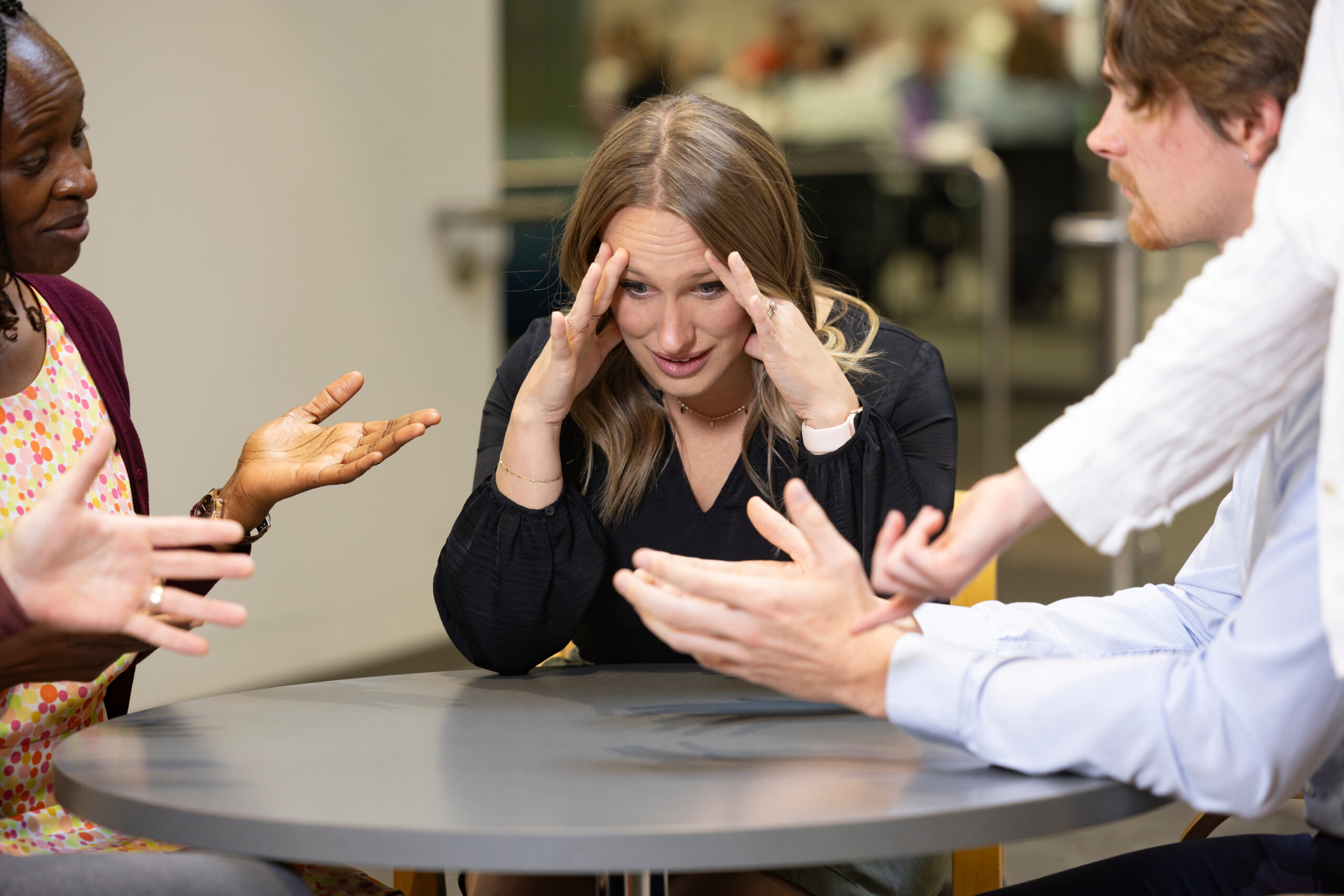 Woman with her hands on her temples listening to people around a table talking and gesturing toward her with their hands.