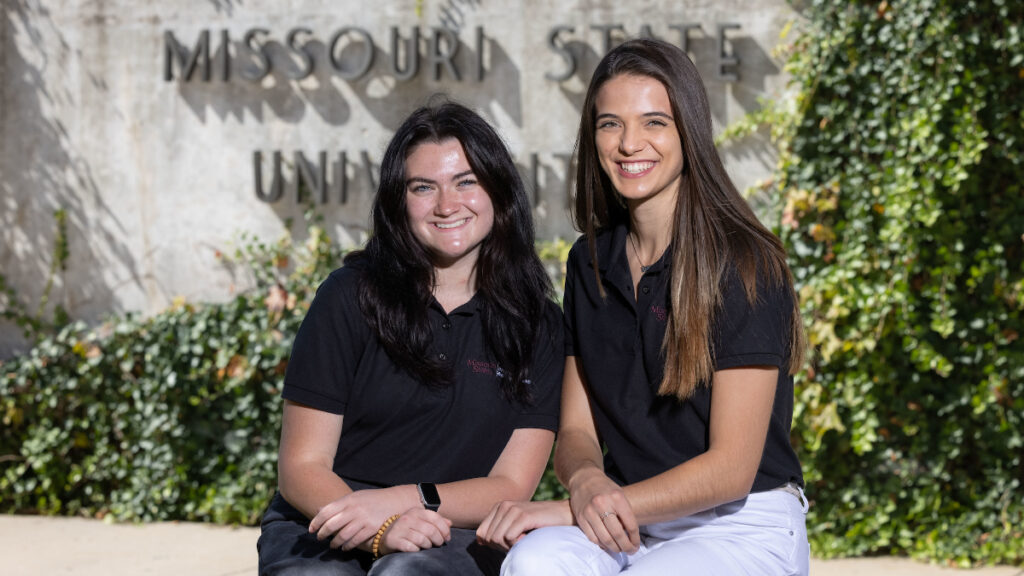 Two female students sit in front of Legacy Wall.