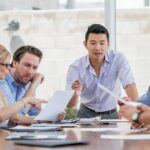Asian businessman at the head of a conference table speaking to a group of employees who are all looking very thoughtfully at several documents on the table and in front of them.