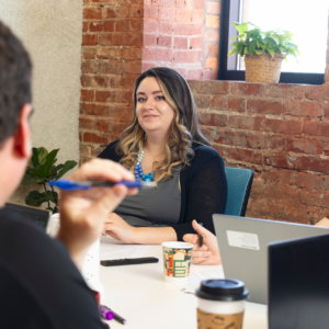 Female business professional looking across a conference table at a man seated across from her holding a pen. Another woman is seated next to her.