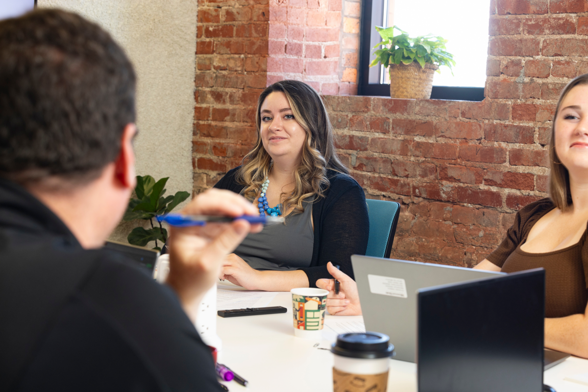 Female business professional looking across a conference table at a man seated across from her holding a pen. Another woman is seated next to her.