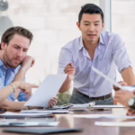 Asian businessman at the head of a conference table speaking to a group of employees who are all looking very thoughtfully at several documents on the table and in front of them.