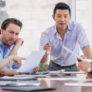 Asian businessman at the head of a conference table speaking to a group of employees who are all looking very thoughtfully at several documents on the table and in front of them.