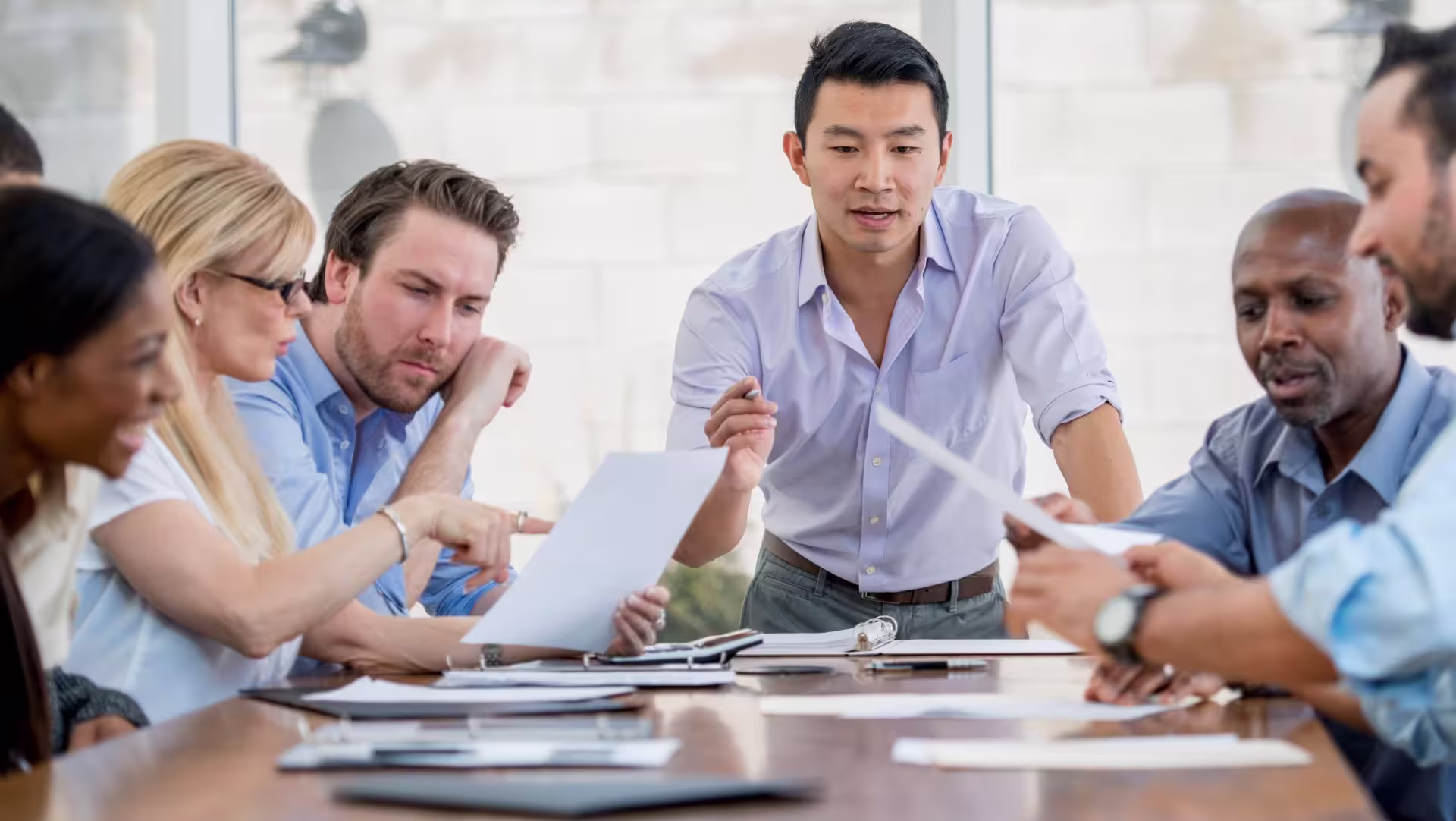 Asian businessman at the head of a conference table speaking to a group of employees who are all looking very thoughtfully at several documents on the table and in front of them.