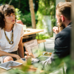 Man talking to a woman at an outdoor table on a patio.