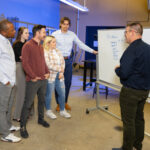 Supervisor speaking to a group of employees in front of a white board of written information.