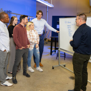 Supervisor speaking to a group of employees in front of a white board of written information.