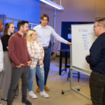 Supervisor speaking to a group of employees in front of a white board of written information.