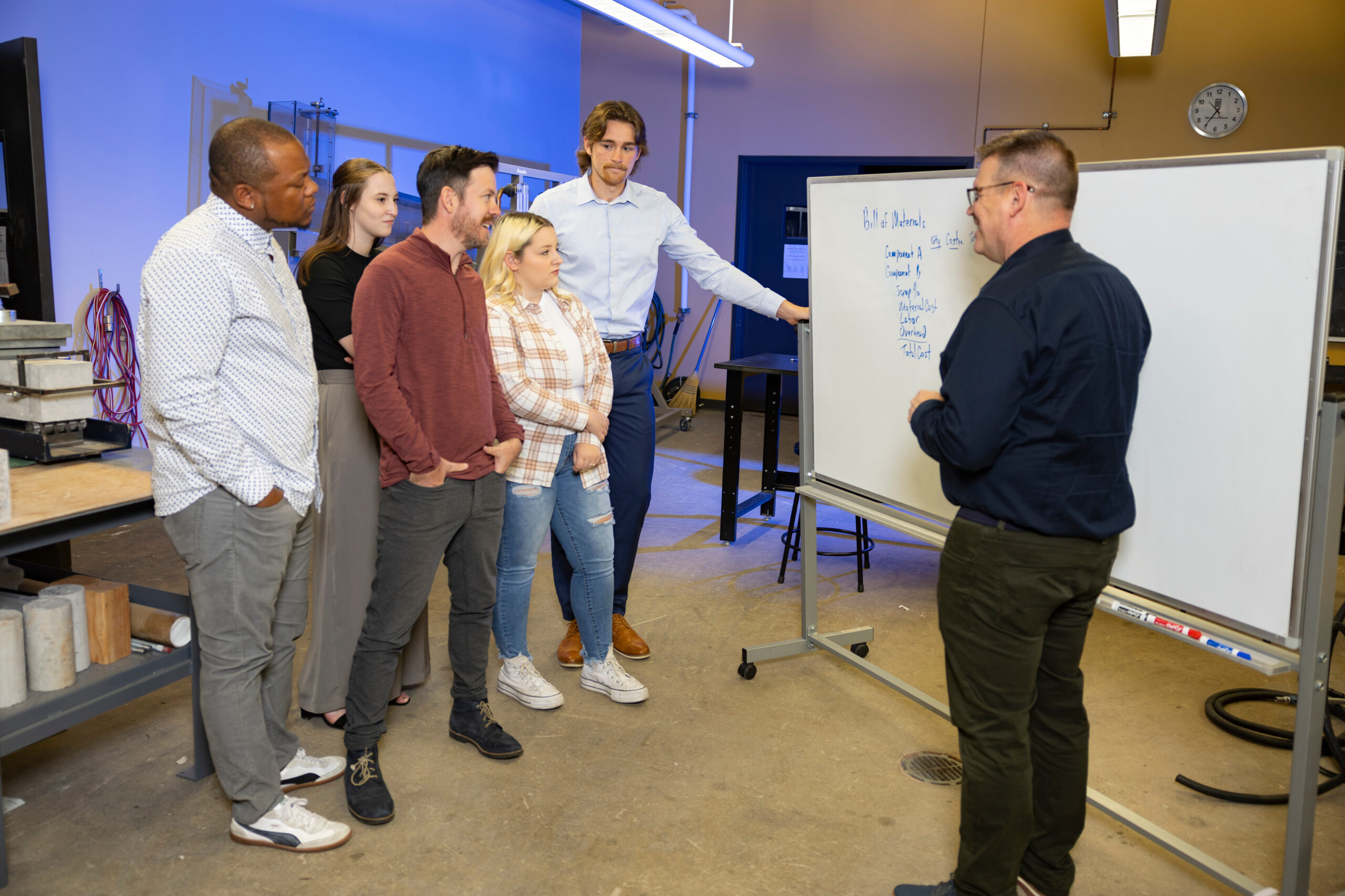 Supervisor speaking to a group of employees in front of a white board of written information.
