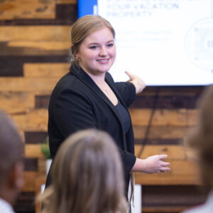 Blonde, Caucasian woman gesturing toward a presentation on a televsion screen in front of a group of seated people.