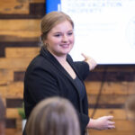 Blonde, Caucasian woman gesturing toward a presentation on a televsion screen in front of a group of seated people.