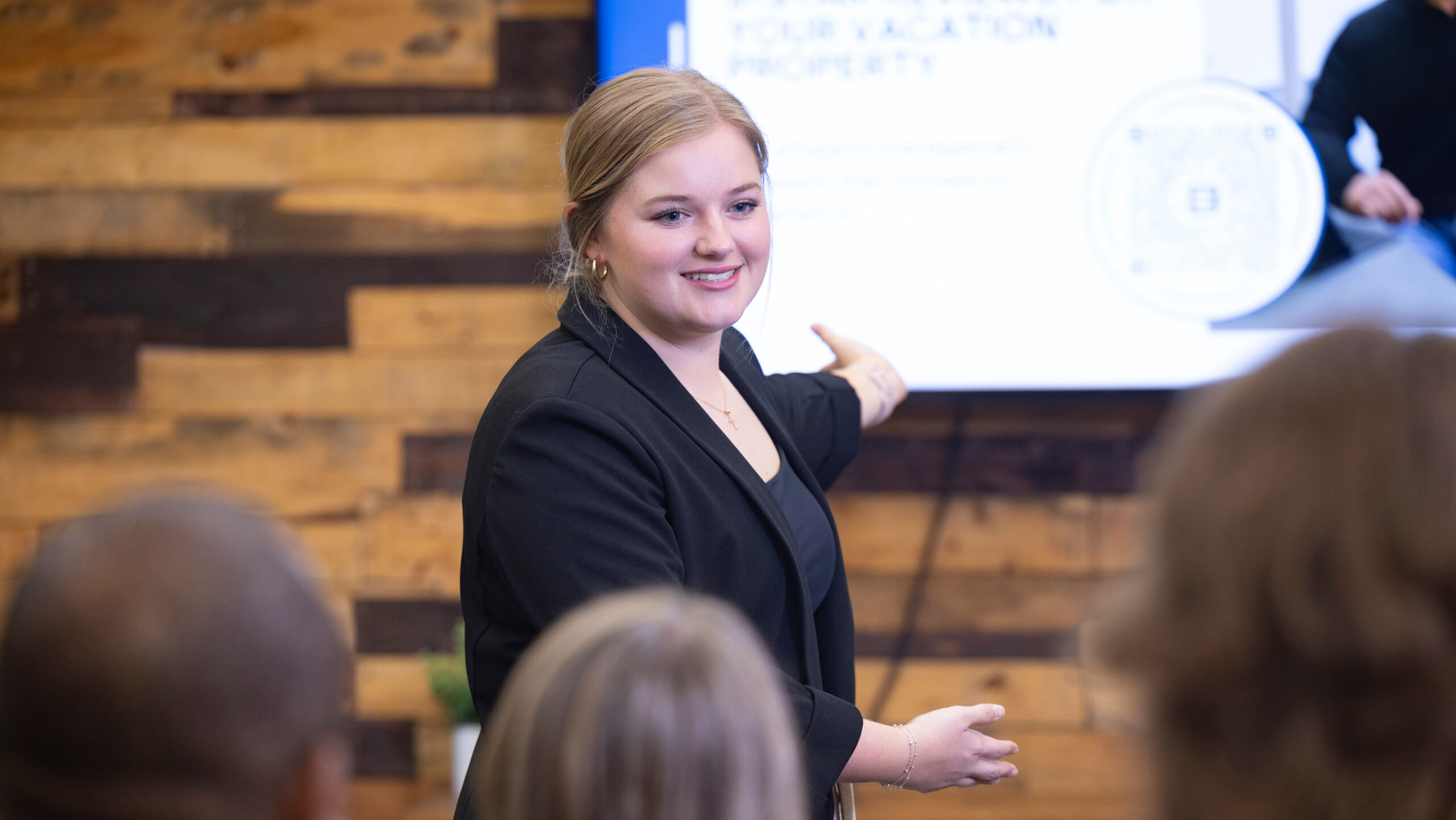 Blonde, Caucasian woman gesturing toward a presentation on a televsion screen in front of a group of seated people.