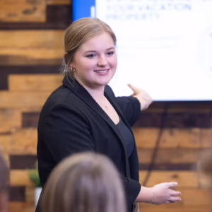 Blonde, Caucasian woman gesturing toward a presentation on a televsion screen in front of a group of seated people.