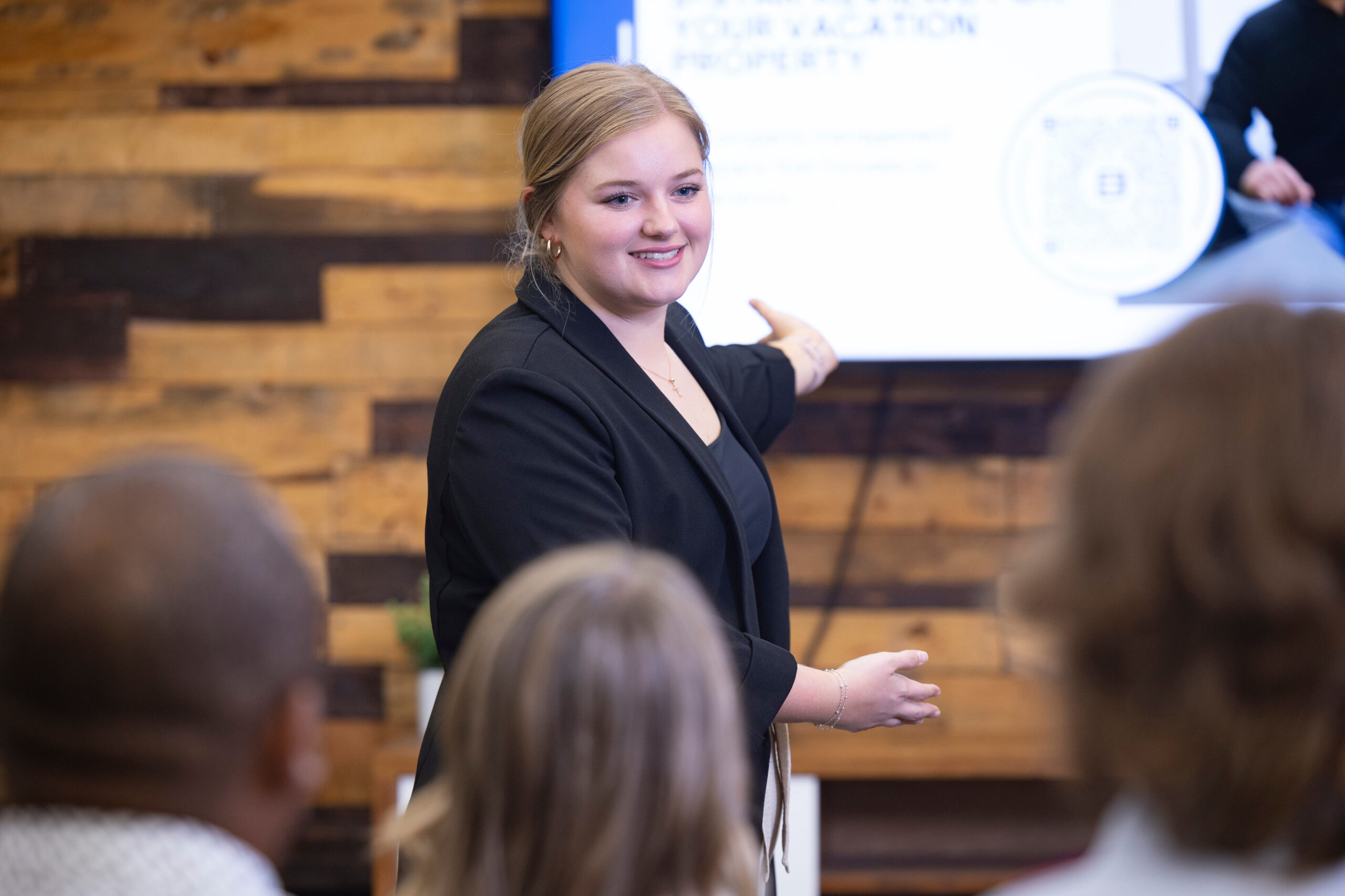 Blonde, Caucasian woman gesturing toward a presentation on a televsion screen in front of a group of seated people.