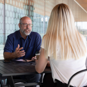 Bald, Caucasian man with a bear in a blue shirt sitting across a table gesturing toward a blonde, Caucasian women in conversation.