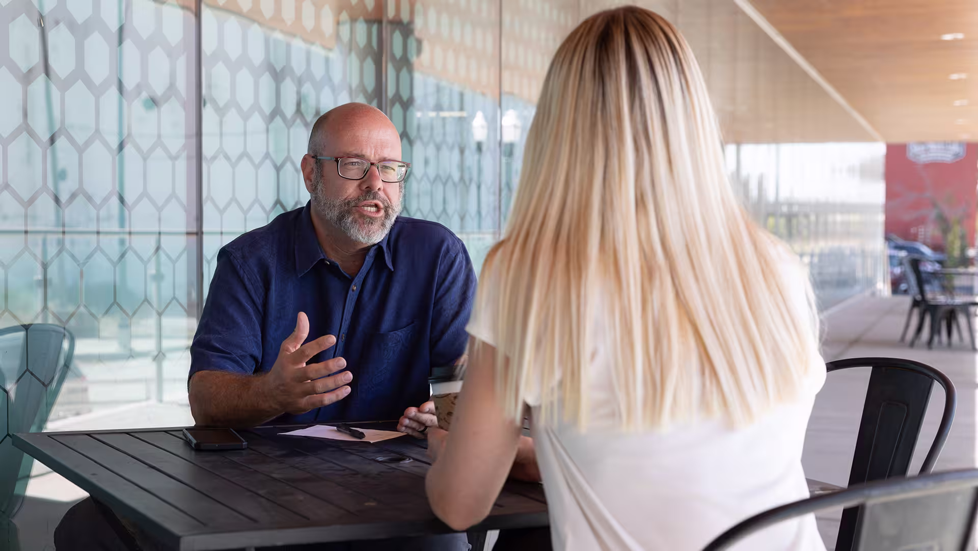 Bald, Caucasian man with a bear in a blue shirt sitting across a table gesturing toward a blonde, Caucasian women in conversation.