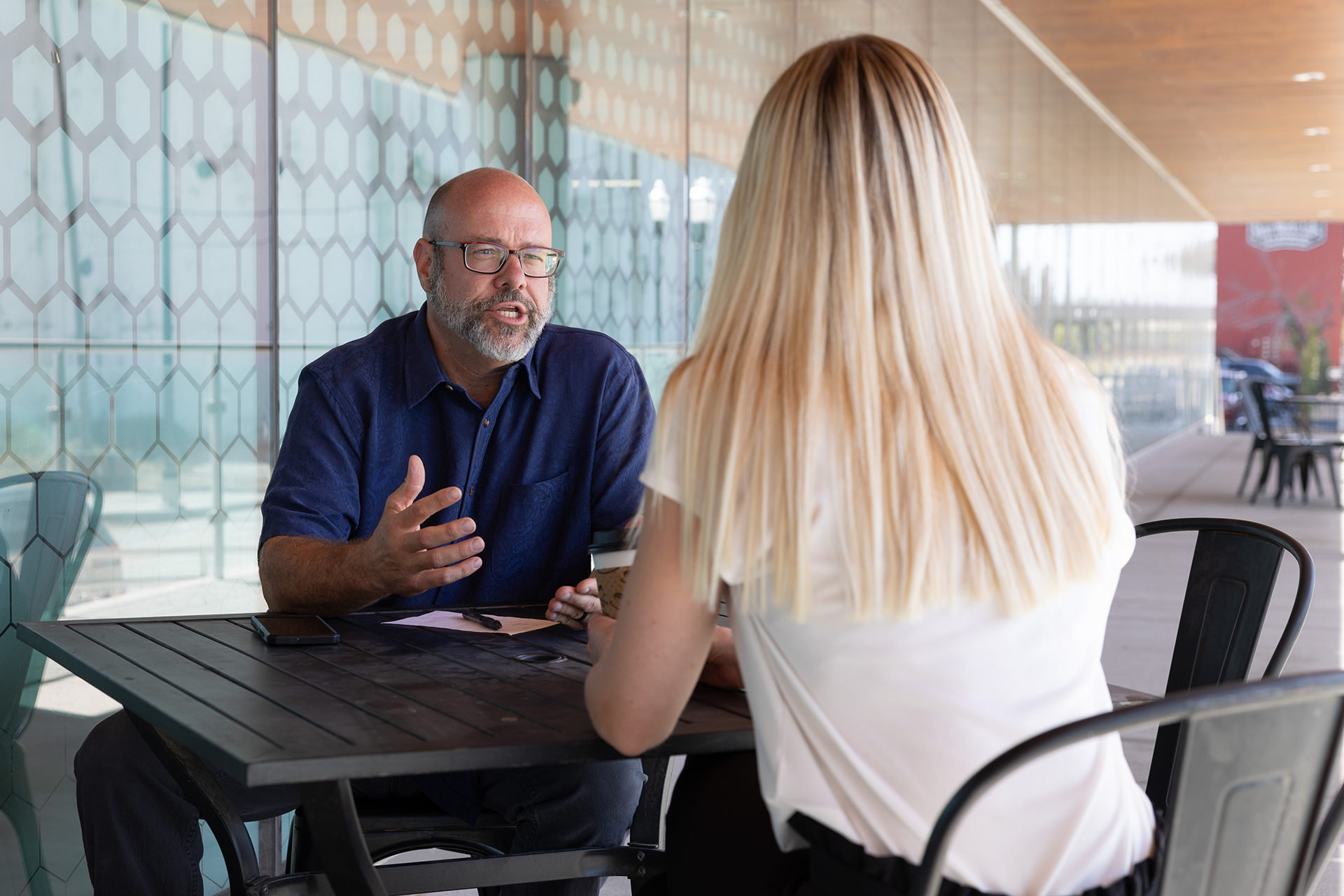 Bald, Caucasian man with a bear in a blue shirt sitting across a table gesturing toward a blonde, Caucasian women in conversation.