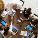 Group of business people sitting around a table with their laptops. Two of the people are shaking hands across the table.