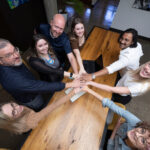 Group of business professionals gathered around a conference table putting their hands in a circle, stacked on top of each other as a team.