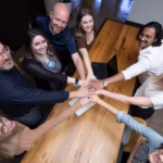 Group of business professionals gathered around a conference table putting their hands in a circle, stacked on top of each other as a team.