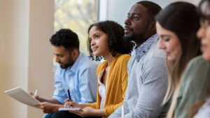 A group of business people sit in a row in a training class. They look at an unseen speaker as they concentrate on his lecture.