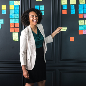 Woman standing in front of board of Post-it notes while man and woman watch her.