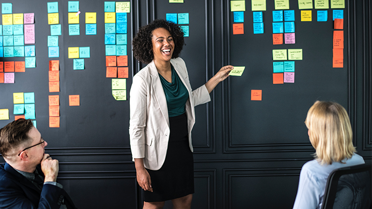 Woman standing in front of board of Post-it notes while man and woman watch her.