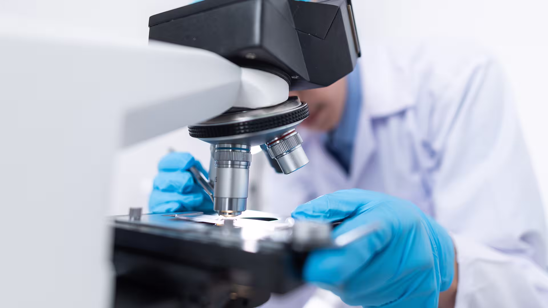 Man in white lab coat and blue rubber gloves, viewing something under a microscope.